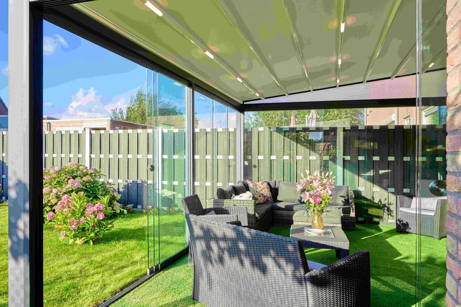 Enclosed glass veranda with a retractable fabric roof and lighting, furnished with dark rattan outdoor furniture, overlooking a green lawn.