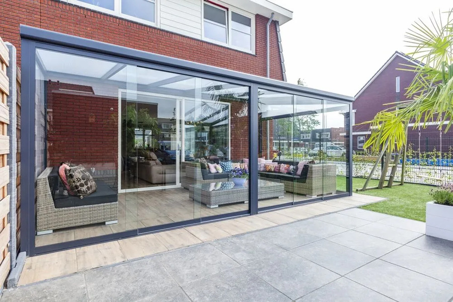 Fully enclosed glass veranda with a dark frame, furnished as an outdoor living room with grey rattan sofas, extending onto a patio.