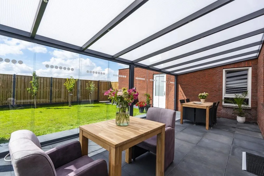 Partially enclosed veranda with a clear, ribbed roof and glass sliding walls, furnishing an outdoor dining area adjacent to a red brick house and garden.