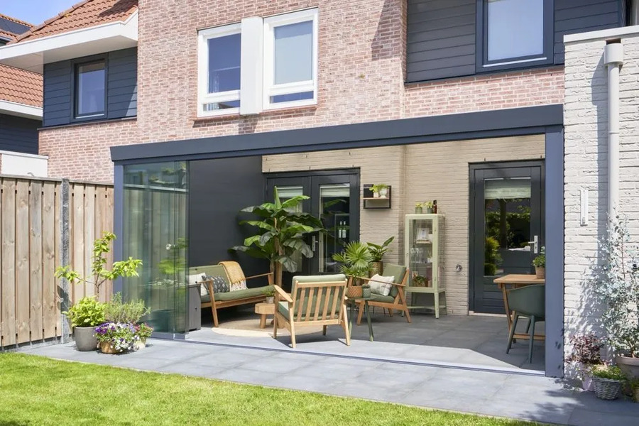 Open-sided veranda structure attached to a house, featuring an indoor/outdoor living space with wood and wicker furniture and plants.